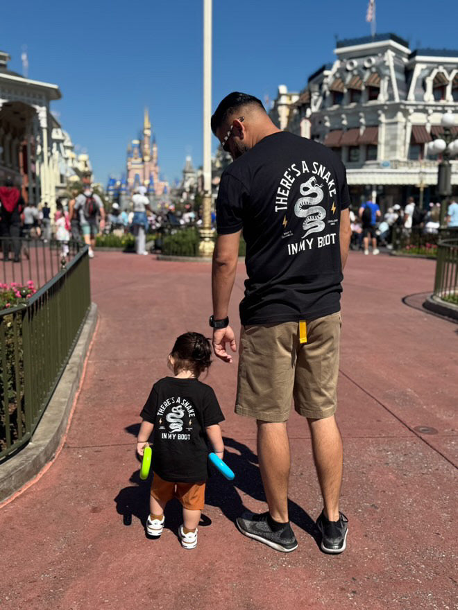 A dad in a black Snake in My Boot tee walks hand in hand with his toddler down Main Street