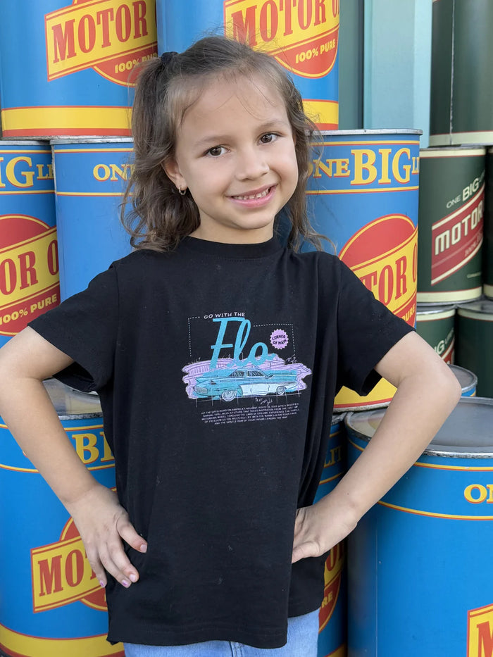 Girl in a black Flo's V8 Cafe inspired kids tee posing in front of oil cans